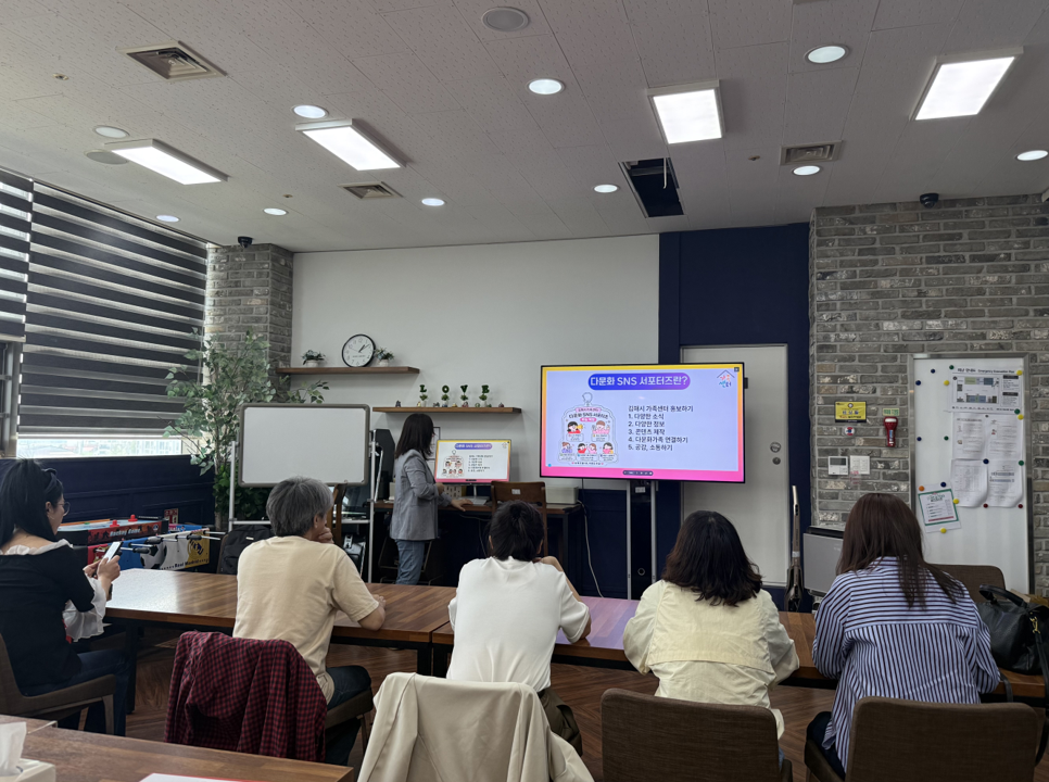 [이미지 내용 및 텍스트 전문] 강의실에서 '다문화 SNS 서포터즈' 교육이 진행 중인 모습. 중앙의 대형 모니터에는 분홍색 배경의 발표 자료가 띄워져 있으며, 강사가 이를 설명하고 있다. 앞에는 5명의 참가자가 테이블에 앉아 경청하고 있다.  [모니터 내 슬라이드 텍스트 내용]  제목: 다문화 SNS 서포터즈란?  내용: 김해시 가족센터 홍보하기  다양한 소식  유익한 정보  콘텐츠 제작  다문화 가족 연결하기  공유, 소통하기