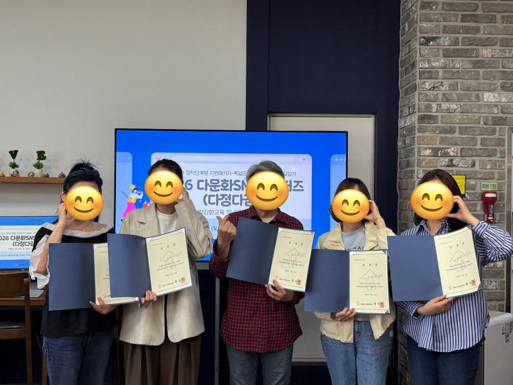 [이미지 내용 및 텍스트 전문] '2026 다문화 SNS 서포터즈' 위촉식 기념 촬영 사진. 5명의 위촉자가 파란색 표지의 위촉장을 펼쳐 들고 나란히 서 있다. 배경의 대형 모니터에는 위촉식 관련 슬라이드가 띄워져 있으며, 참석자들은 얼굴이 미소 짓는 이모티콘으로 가려진 채 손하트 등의 포즈를 취하고 있다.  [모니터 및 위촉장 주요 텍스트]  배경 모니터: 2026 다문화 SNS 서포터즈 (다정다감) 역량강화교육 및 발대식  위촉장 내용: 위촉장, 김해시가족센터. 귀하를 김해시가족센터에서 운영하는 '다문화 SNS 서포터즈' 위원(단원)으로 위촉함. 2026년 4월 24일. 김해시가족센터장.